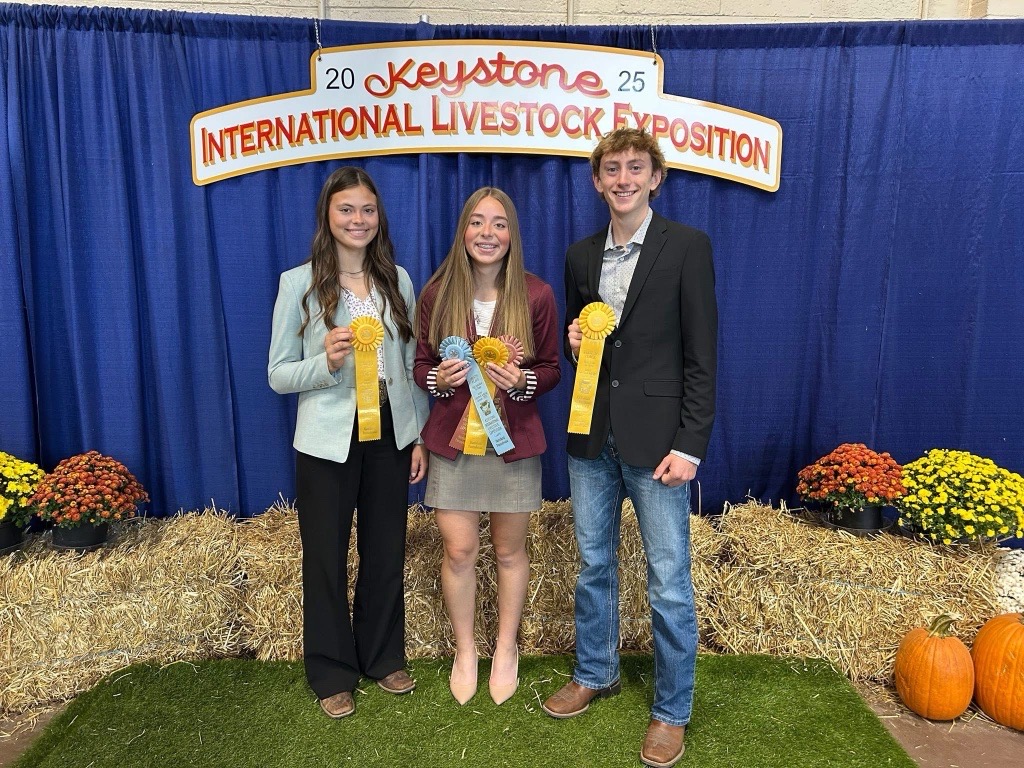 Two teen girls and one teen boy holding up their ribbons in front of a sign that says 2025 Keystone International Livestock Exposition.